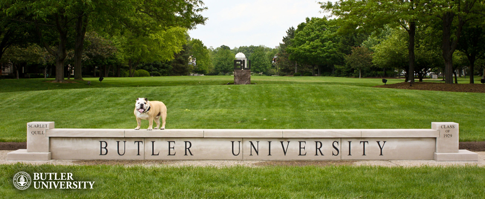 A bulldog stands on a large stone wall engraved with the words "Butler University". A green field, framed by trees, can be seen in the background.