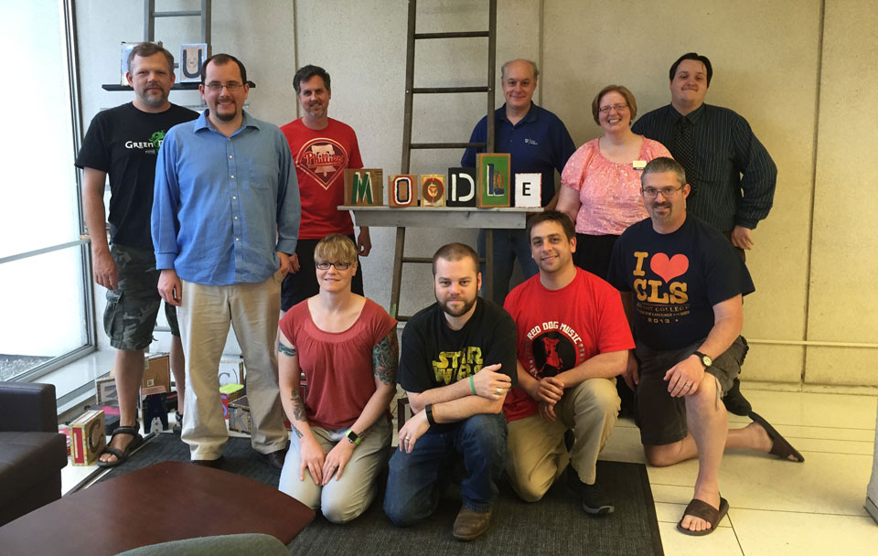 The ten people who attended Moodle Hack/Doc Fest stand on either side of stylized block letters that spell out the word "Moodle".. The 