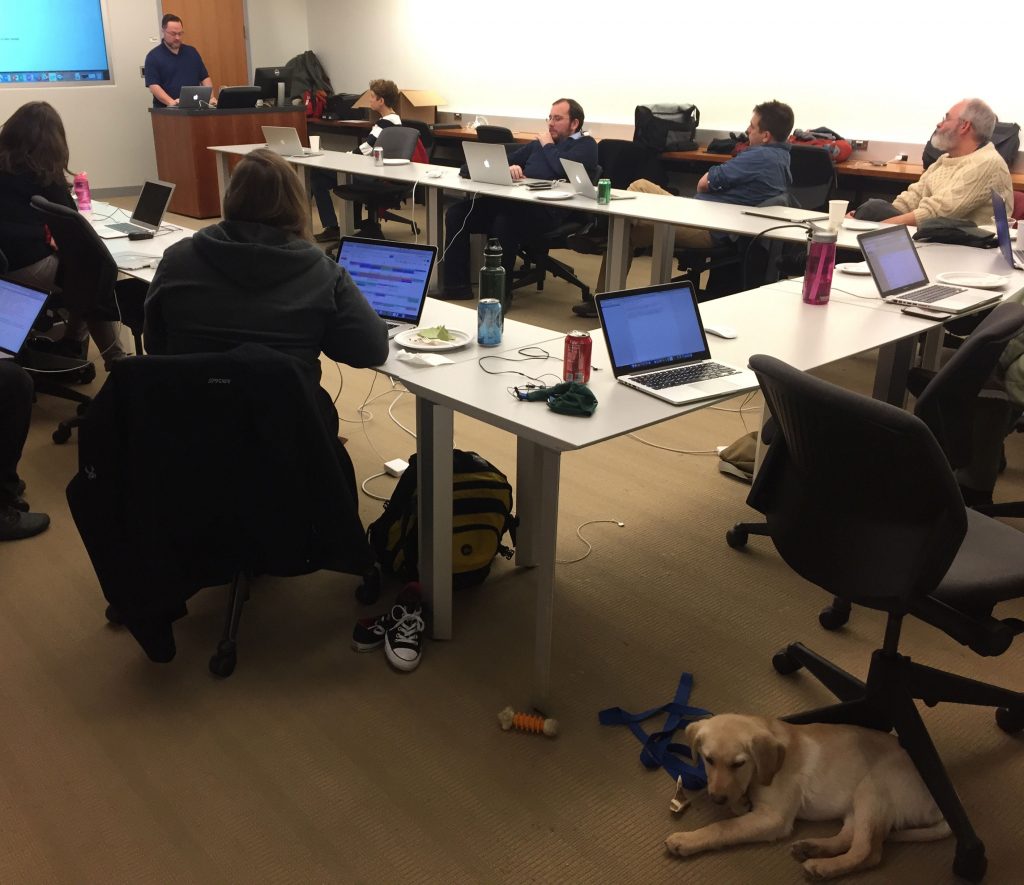 Attendees at Hack/Doc sit around a U-shaped table while a male presenter talks. A small Yellow Labrador puppy is laying down in the foreground.